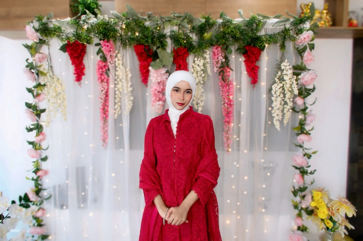 A woman in a striking red dress stands in front of a lush floral backdrop, showcasing a vibrant scene.