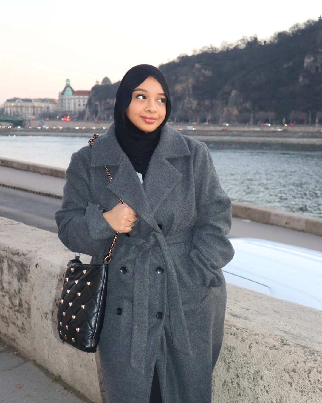 A woman in a stylish coat and scarf stands for a photo, looking confidently at the camera with a warm smile.