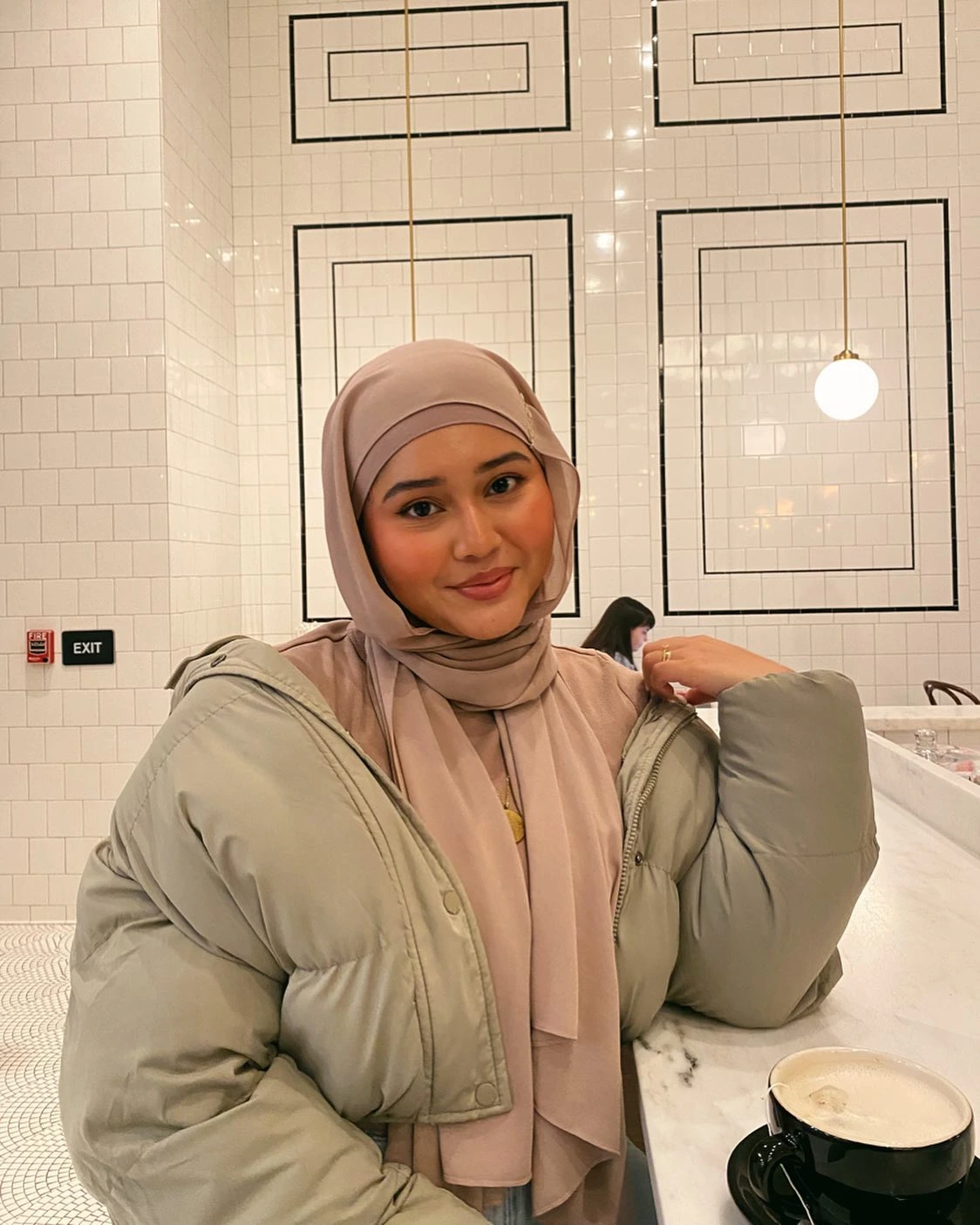 A woman in a hijab sits at a table, enjoying a cup of coffee in a cozy setting