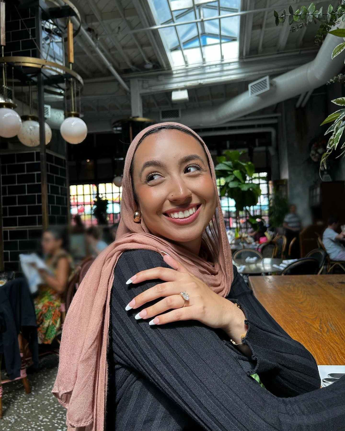 A smiling woman wearing a hijab sits at a table, radiating positivity and warmth in her expression.