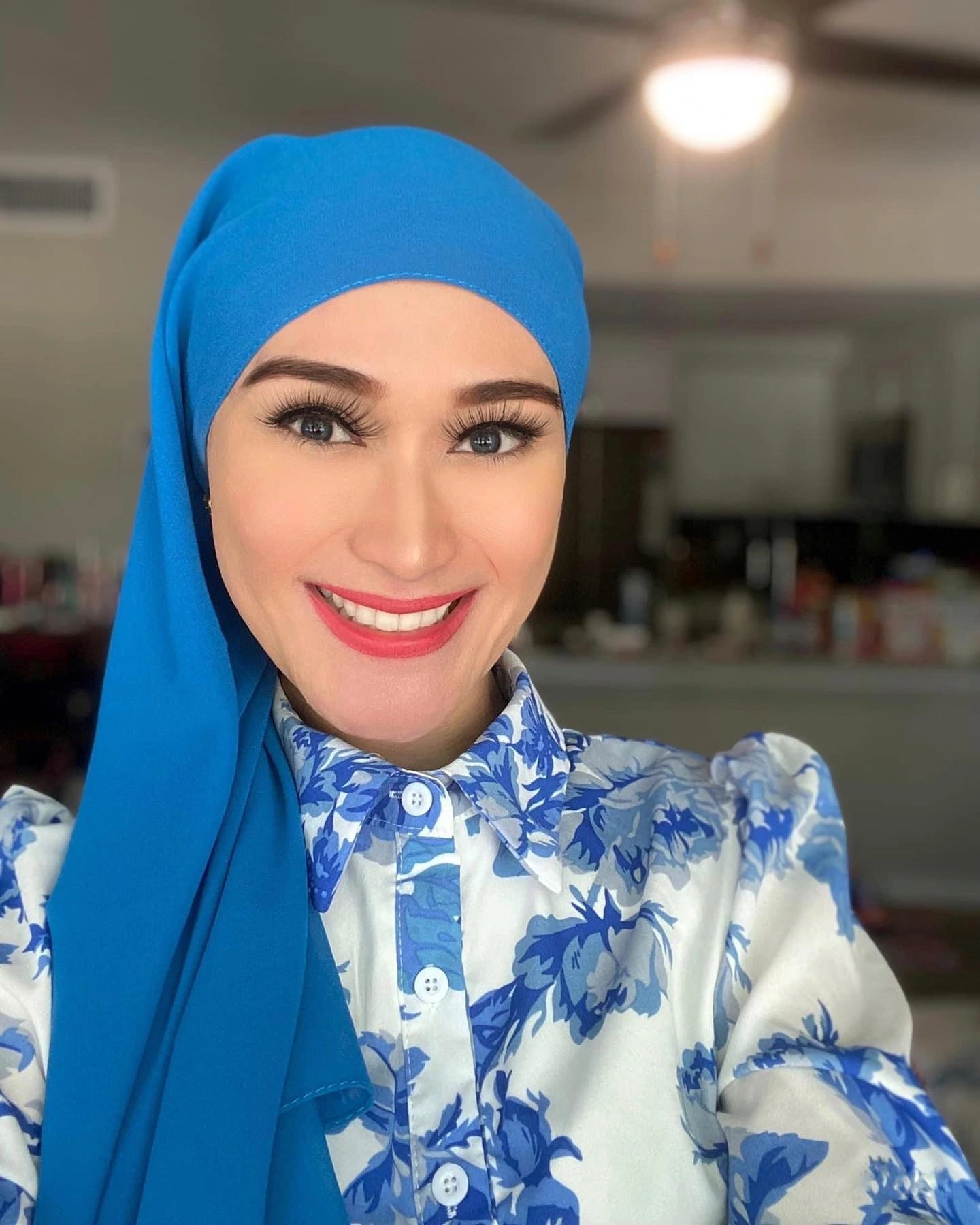 A woman in a blue headscarf and matching blue shirt, smiling softly against a neutral background.